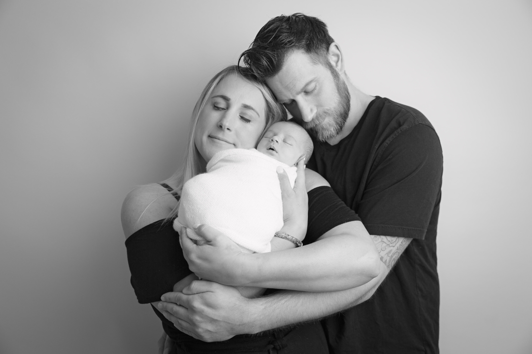 black and white photo of new parents posed holding their wrapped baby in a studio setting against a backdrop, parents cuddled in together and posed with their baby by hayley morris photography